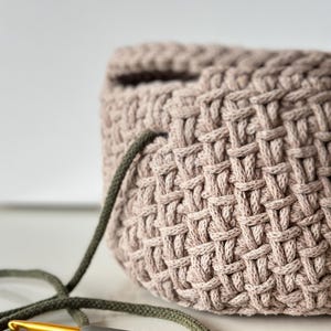 May include: A close-up of a crocheted basket made with a light brown yarn. A green and gold crochet hook is lying on a white surface next to the basket.
