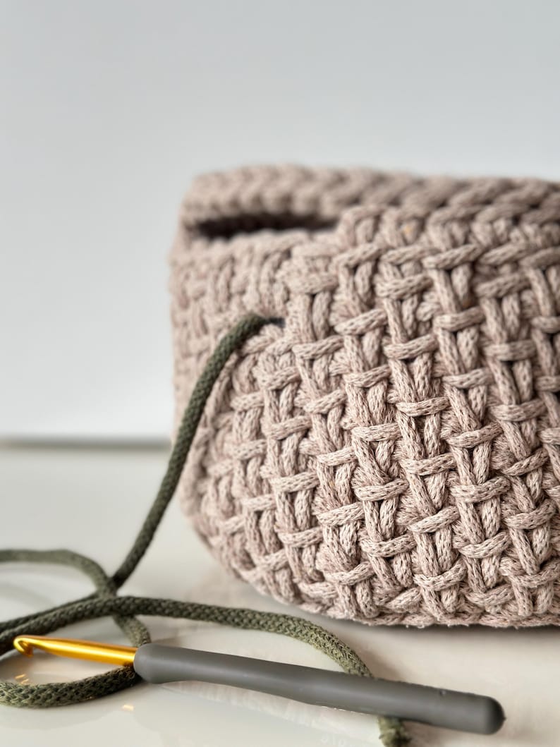 May include: A close-up of a crocheted basket made with a light brown yarn. A green and gold crochet hook is lying on a white surface next to the basket.
