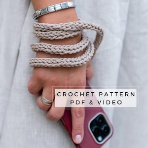 Puede incluir: Primer plano de una mano sosteniendo una funda de teléfono roja con una correa de teléfono de ganchillo. El texto "CROCHET PATTERN PDF & VIDEO" se muestra en la imagen.