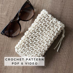 Könnte beinhalten: Eine Sonnenbrille mit Schildpattmuster liegt neben einem cremefarbenen, gehäkelten Brillenetui mit Kordelzug. Das Etui hat ein strukturiertes, gewebtes Aussehen. Text auf dem Bild: CROCHET PATTERN PDF & VIDEO.