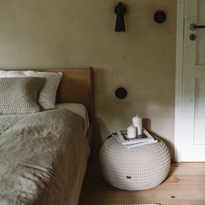 K&ouml;nnte beinhalten: Ein Schlafzimmer mit einem Bett mit einer beigefarbenen Bettw&auml;sche aus Leinen und einem gestrickten &Uuml;berwurfkissen. Ein wei&szlig;er geh&auml;kelter Hocker mit zwei wei&szlig;en Kerzen und einem Magazin darauf steht neben dem Bett. Ein hellgrauer gestrickter Teppich liegt auf dem Boden.