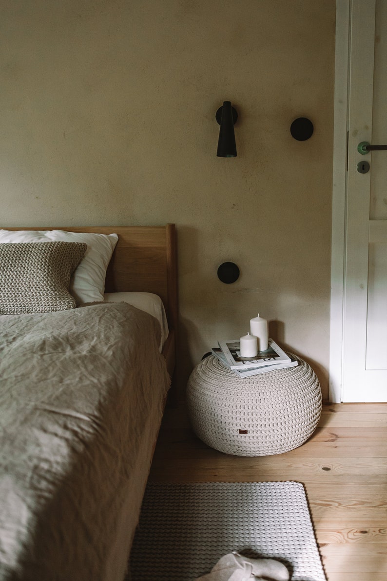 K&ouml;nnte beinhalten: Ein Schlafzimmer mit einem Bett mit einer beigefarbenen Bettw&auml;sche aus Leinen und einem gestrickten &Uuml;berwurfkissen. Ein wei&szlig;er geh&auml;kelter Hocker mit zwei wei&szlig;en Kerzen und einem Magazin darauf steht neben dem Bett. Ein hellgrauer gestrickter Teppich liegt auf dem Boden.