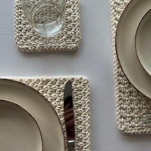 May include: A neutral-toned, crocheted placemat and coaster set. The placemats are rectangular, and the coaster is square. A clear glass with water sits on the coaster. Ceramic plates and silverware are also visible, suggesting a dining setting.