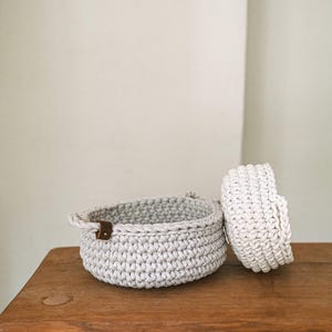 以下が含まれることがあります： 木製のテーブルの上に、2つの編みかごが置かれています。1つは他方よりも大きく、どちらも薄いグレーの色をしています。