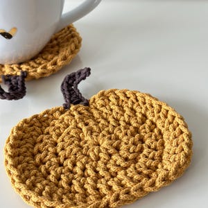 May include: A crocheted pumpkin-shaped coaster in a mustard yellow color with a brown stem. The coaster is made with a textured stitch pattern.