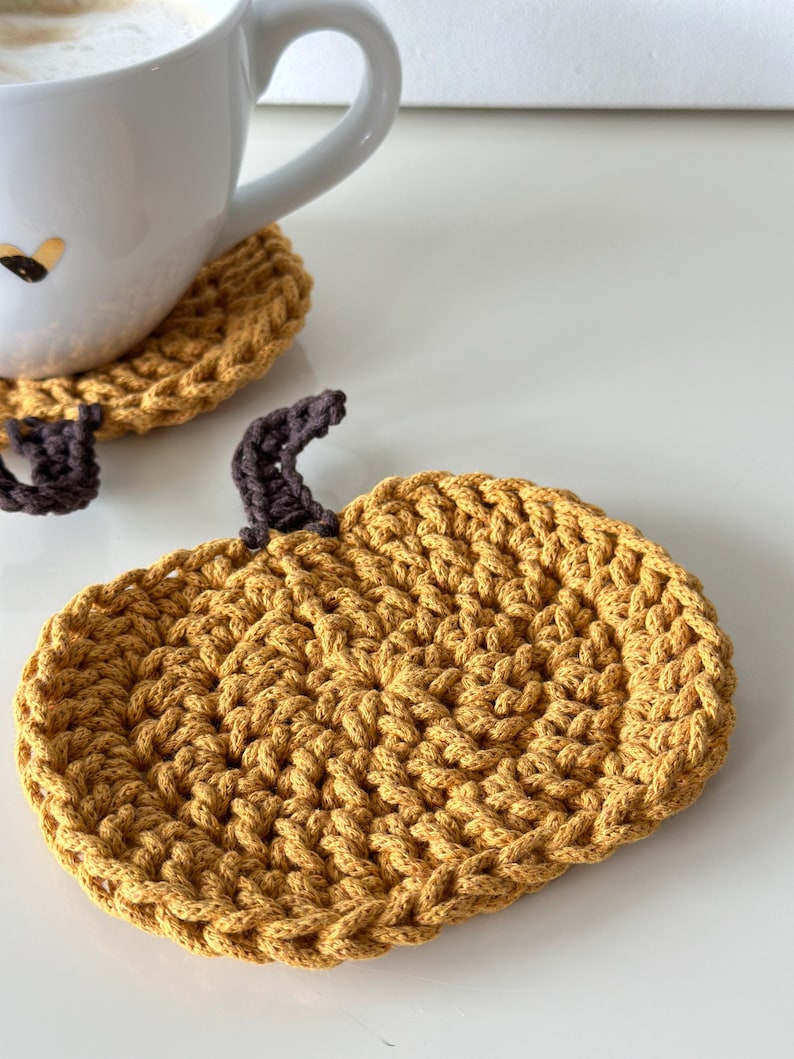 May include: A crocheted pumpkin-shaped coaster in a mustard yellow color with a brown stem. The coaster is made with a textured stitch pattern.