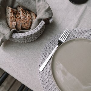 May include: A gray crocheted placemat with a gray ceramic plate and a silver fork on top. The plate is empty. A gray crocheted bowl with a linen cloth inside is filled with slices of bread.