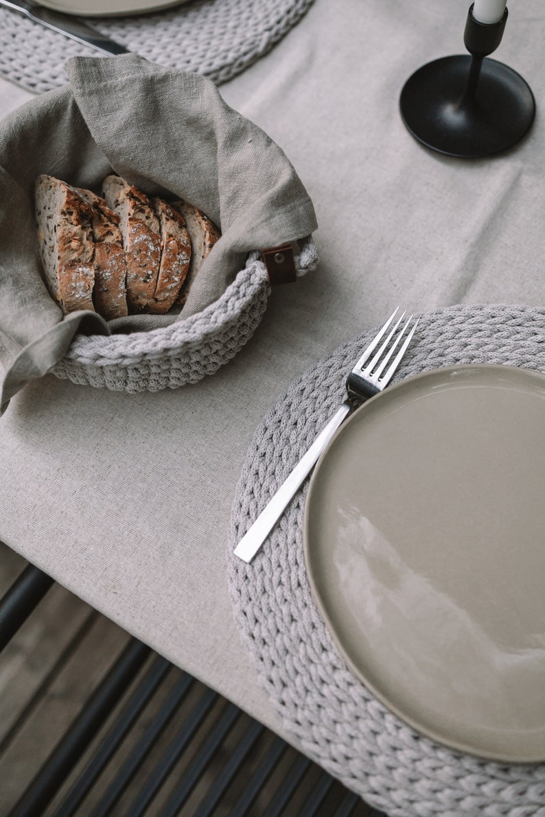 May include: A gray crocheted placemat with a gray ceramic plate and a silver fork on top. The plate is empty. A gray crocheted bowl with a linen cloth inside is filled with slices of bread.