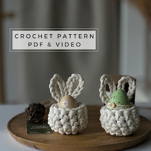 Op de afbeelding: Twee gebreide paaseierenhouders in de vorm van een konijn met een bruin ei en een groen gevlekt ei erin. De houders zijn gemaakt van beige garen en hebben witte bloemenaccenten. De tekst "CROCHET PATTERN PDF & VIDEO" staat bovenaan de afbeelding.