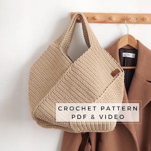 May include: A beige crocheted tote bag hanging on a wooden rack with a brown coat hanging next to it. The text "CROCHET PATTERN PDF & VIDEO" is printed below the bag.