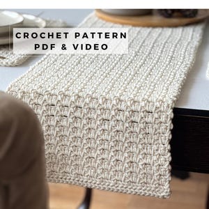 May include: A cream-colored crocheted table runner featuring a textured, repeating stitch pattern. The image includes the text CROCHET PATTERN PDF & VIDEO overlaid in a white box. The runner is displayed on a dark surface with a plate visible.