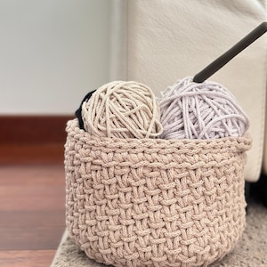 May include: A beige crocheted basket with two balls of yarn inside. One ball of yarn is beige and the other is light purple.