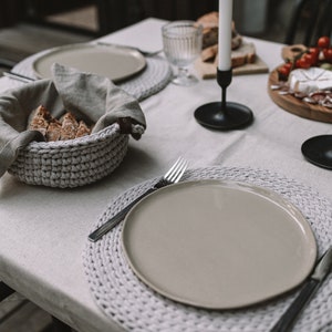 May include: A white tablecloth with a crocheted placemat, a beige plate, and a silver fork. A crocheted basket with bread is on the table. A black candle holder with a white candle is in the background.