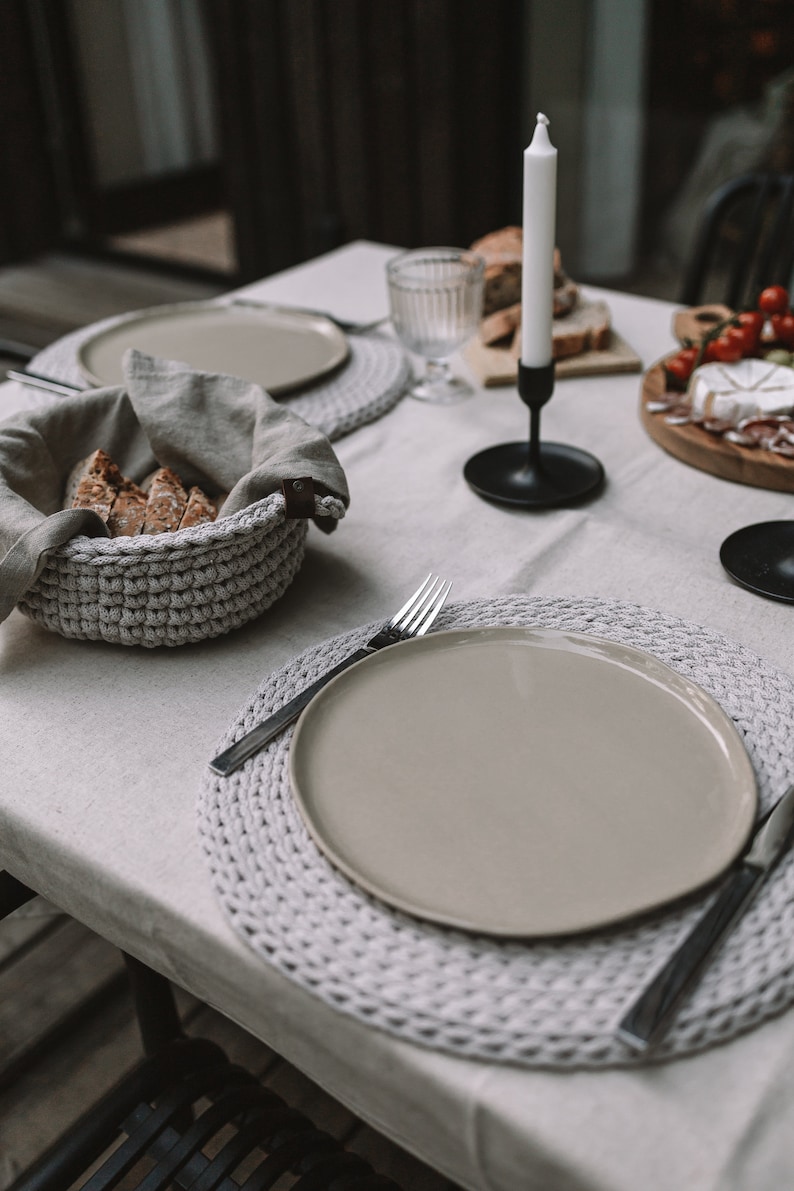 May include: A white tablecloth with a crocheted placemat, a beige plate, and a silver fork. A crocheted basket with bread is on the table. A black candle holder with a white candle is in the background.