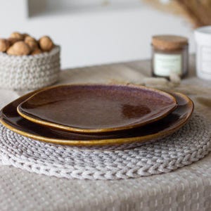 May include: Two stacked ceramic plates with a glossy, dark brown finish and a subtle speckled texture. The plates feature slightly irregular, rounded square edges and a smooth, reflective surface that catches the light, creating depth.