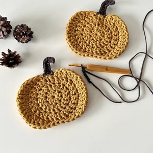 May include: Two crocheted pumpkin coasters in a mustard yellow color with brown stems. The coasters are on a white surface with a crochet hook and yarn.