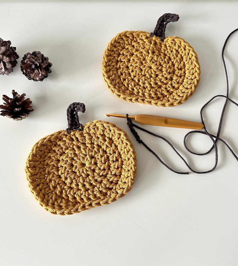 May include: Two crocheted pumpkin coasters in a mustard yellow color with brown stems. The coasters are on a white surface with a crochet hook and yarn.