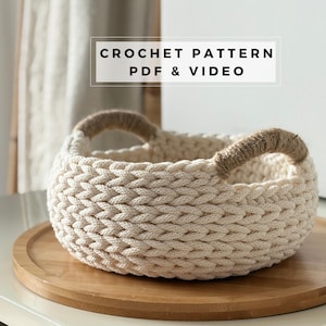 Puede incluir: Una cesta redonda de ganchillo de color crema con asas de cuerda. La cesta está sobre una bandeja de madera. El texto "CROCHET PATTERN PDF & VIDEO" se muestra en un cuadro blanco encima de la cesta.