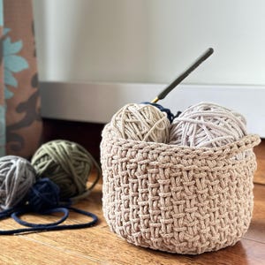 May include: A beige crocheted basket with three balls of yarn inside. Two balls of yarn are beige and one is green. A crochet hook is sticking out of the basket.