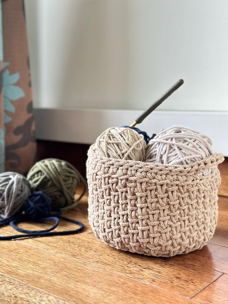 May include: A beige crocheted basket with three balls of yarn inside. Two balls of yarn are beige and one is green. A crochet hook is sticking out of the basket.