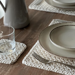 May include: A dining table setting with cream-colored crochet placemats, plates, and a fork. A gray pitcher and a glass with a deer emblem are also on the table. The plates have a light brown rim.