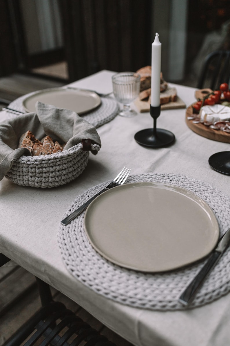 May include: A table setting featuring round, light gray crocheted placemats, a matching crocheted bread basket lined with a beige linen cloth, beige ceramic plates, stainless steel silverware, and a black candle holder with a white candle.
