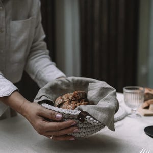 以下が含まれることがあります： 人が、薄い灰色の編み込みのバスケットを、茶色の革の取っ手を持って持っています。バスケットはパンの切れ端でいっぱいです。バスケットは白いテーブルクロスの上に置かれています。