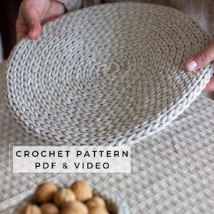 May include: A round, beige crochet placemat featuring a dense, spiraling stitch pattern. The text CROCHET PATTERN PDF & VIDEO is overlaid in black on a white rectangular background. The item is made of thick, textured cord-like yarn.