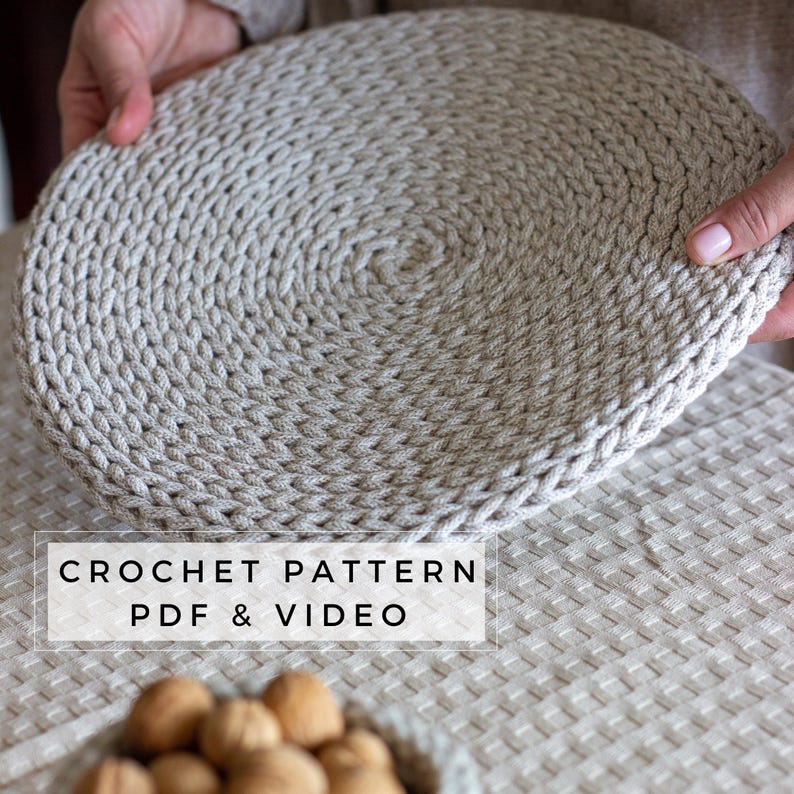 May include: A round, beige crochet placemat featuring a dense, spiraling stitch pattern. The text CROCHET PATTERN PDF & VIDEO is overlaid in black on a white rectangular background. The item is made of thick, textured cord-like yarn.