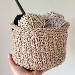 May include: A beige crocheted basket with a woven pattern holds three balls of yarn in beige, light gray, and black.