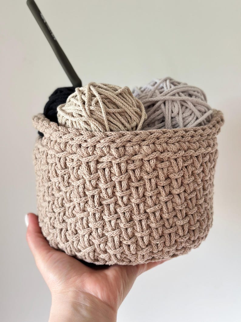 May include: A beige crocheted basket with a woven pattern holds three balls of yarn in beige, light gray, and black.