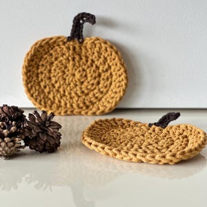 May include: Two crocheted pumpkin coasters in a mustard yellow color with brown stems. The coasters are on a white surface with pine cones.