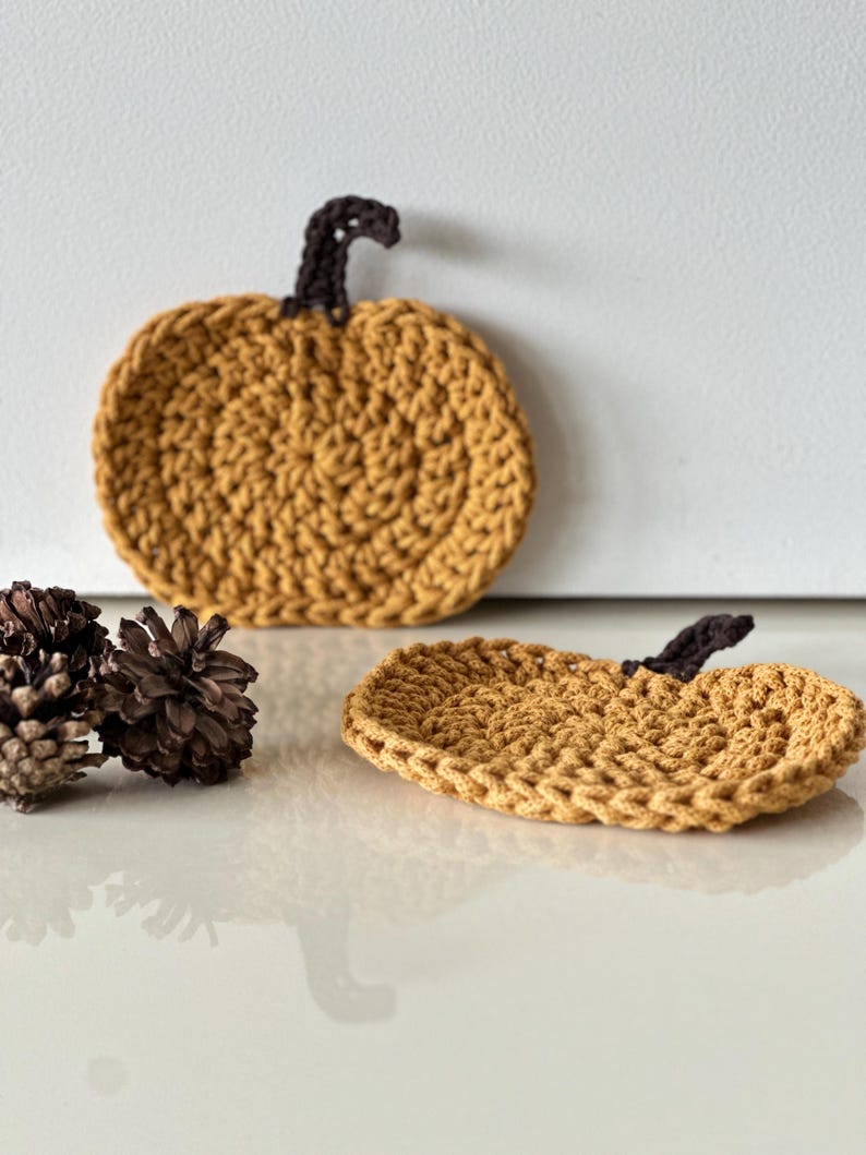 May include: Two crocheted pumpkin coasters in a mustard yellow color with brown stems. The coasters are on a white surface with pine cones.