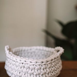 以下が含まれることがあります： 2つの取っ手付きの白い編みかご。かごは空で、木製の表面の上に置かれています。