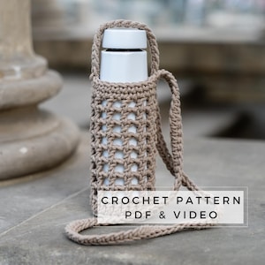 Op de afbeelding: Een gehaakte beige fleshouder met een lange band. De houder is gemaakt met een eenvoudig steekpatroon en heeft een licht open weefsel. De tekst "CROCHET PATTERN PDF & VIDEO" is gedrukt op een wit rechthoek in het onderste midden van de afbeelding.