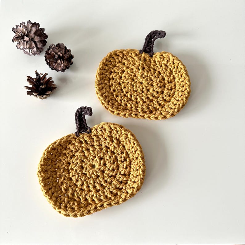 May include: Two crocheted pumpkin-shaped coasters in a mustard yellow color. The coasters have a brown crocheted stem.