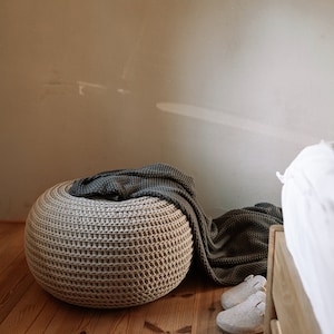 K&ouml;nnte beinhalten: Ein beiger gestrickter Hocker mit einer dunkelgrauen Decke dar&uuml;ber. Der Hocker steht auf einem Holzboden. Neben dem Hocker liegen zwei wei&szlig;e Hausschuhe auf dem Boden.