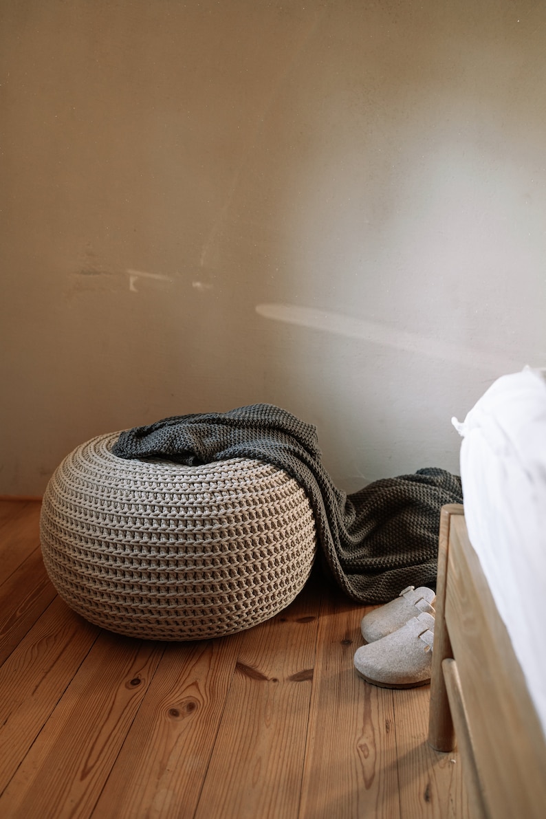 K&ouml;nnte beinhalten: Ein beiger gestrickter Hocker mit einer dunkelgrauen Decke dar&uuml;ber. Der Hocker steht auf einem Holzboden. Neben dem Hocker liegen zwei wei&szlig;e Hausschuhe auf dem Boden.