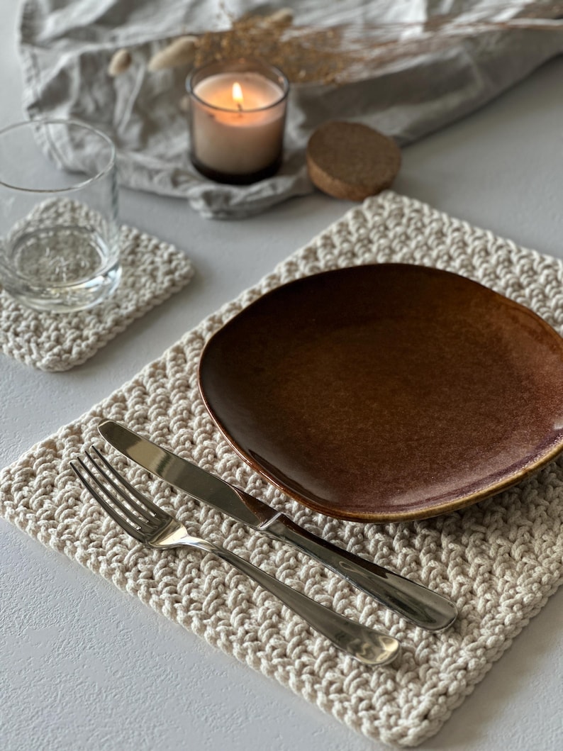 May include: A neutral-toned, crocheted placemat with a brown plate, silverware, and a matching coaster. A lit candle in a glass holder and a clear glass are also present. The scene is set on a light-colored surface, creating a cozy, rustic aesthetic.