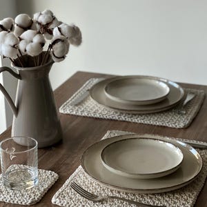 May include: A table setting featuring beige ceramic plates, a glass, and a vase with cotton stems. The plates rest on woven placemats, with silverware arranged beside them. A coaster and a glass with a deer graphic are also visible.