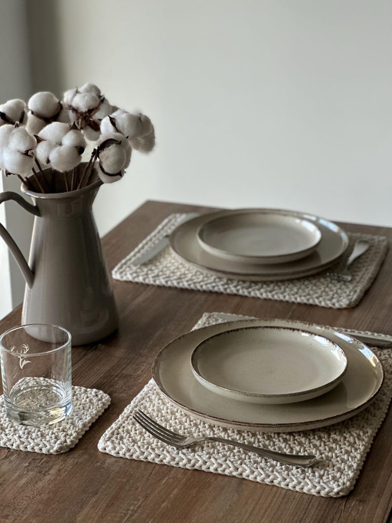 May include: A table setting featuring beige ceramic plates, a glass, and a vase with cotton stems. The plates rest on woven placemats, with silverware arranged beside them. A coaster and a glass with a deer graphic are also visible.