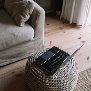 K&ouml;nnte beinhalten: Ein schwarzer Laptop ist ge&ouml;ffnet und liegt auf einem beigen, gestrickten Hocker. Der Hocker steht auf einem hellbraunen Holzboden mit einem gewebten Teppich.