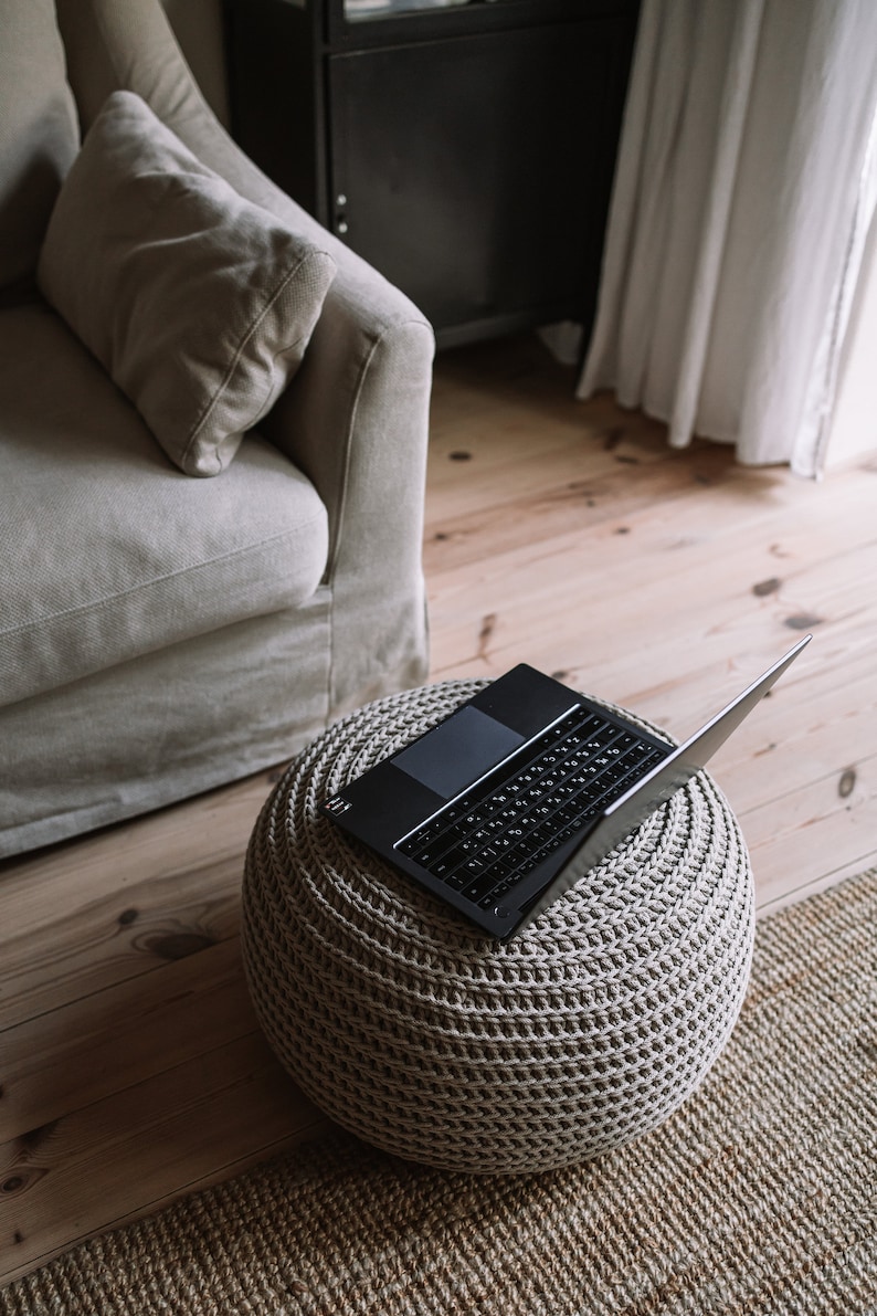 K&ouml;nnte beinhalten: Ein schwarzer Laptop ist ge&ouml;ffnet und liegt auf einem beigen, gestrickten Hocker. Der Hocker steht auf einem hellbraunen Holzboden mit einem gewebten Teppich.