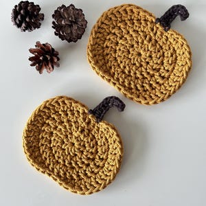 May include: Two crocheted pumpkin-shaped coasters in a mustard yellow color with brown stems. The coasters are on a white surface.