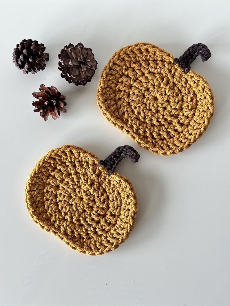 May include: Two crocheted pumpkin-shaped coasters in a mustard yellow color with brown stems. The coasters are on a white surface.