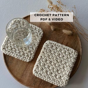 Könnte beinhalten: Zwei beige gehäkelte Untersetzer, einer mit einem Wasserglas, auf einem Holztablett präsentiert. Die Untersetzer haben ein strukturiertes, gewebtes Muster. Der Text auf dem Bild lautet "CROCHET PATTERN PDF & VIDEO."