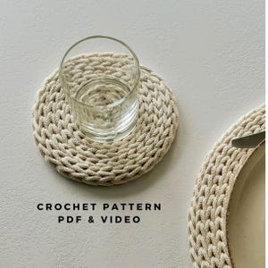 May include: A glass of water rests on a beige, round, crocheted coaster with a braided texture. The text "CROCHET PATTERN PDF & VIDEO" is below the coaster. A matching crocheted placemat is partially visible. The coaster is approximately 10 cm in diameter.