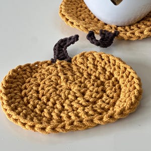 May include: A crocheted pumpkin-shaped coaster in a mustard yellow color with a brown crocheted stem.