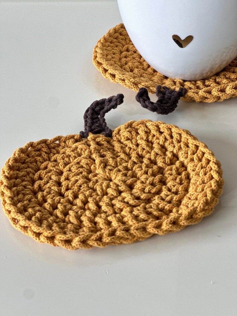May include: A crocheted pumpkin-shaped coaster in a mustard yellow color with a brown crocheted stem.