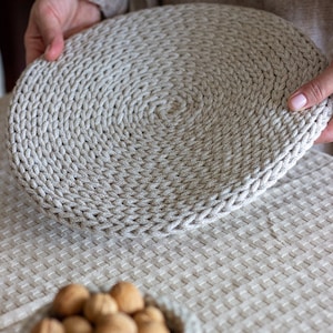 May include: A close-up of a beige crocheted round placemat being held by a hand. The placemat is made of thick rope and has a circular pattern. The placemat is sitting on a beige tablecloth.